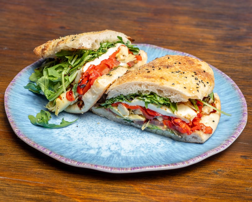Two halves of a focaccia sandwich filled with arugula, roasted red peppers, and cheese on a blue plate.