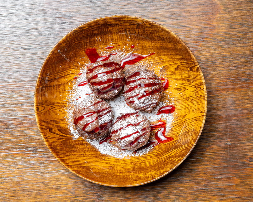 Four chocolate cream puffs with raspberry drizzle and powdered sugar on a wooden plate.