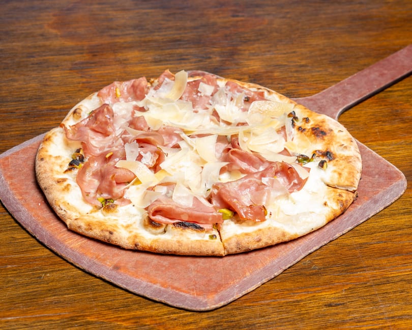 A prosciutto and parmesan pizza on a wooden pizza peel, resting on a dark wooden table.