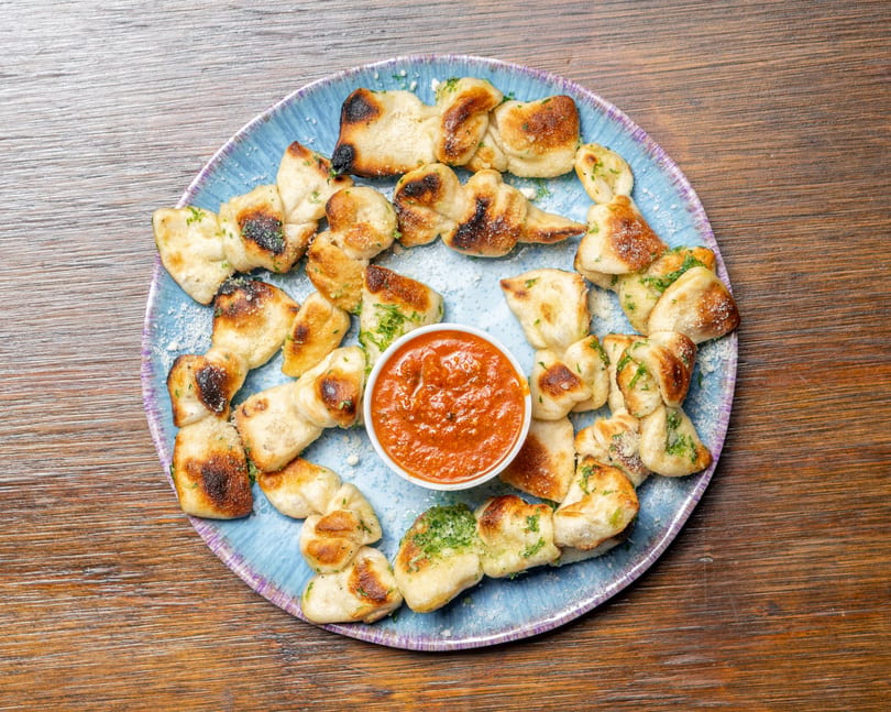 Garlic knots with parmesan and parsley, arranged around a small bowl of marinara sauce on a blue plate.