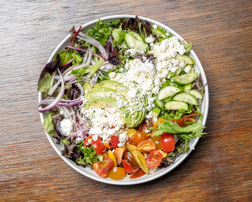 A vibrant salad with mixed greens, red onions, avocado, cucumbers, cherry tomatoes, and feta cheese.