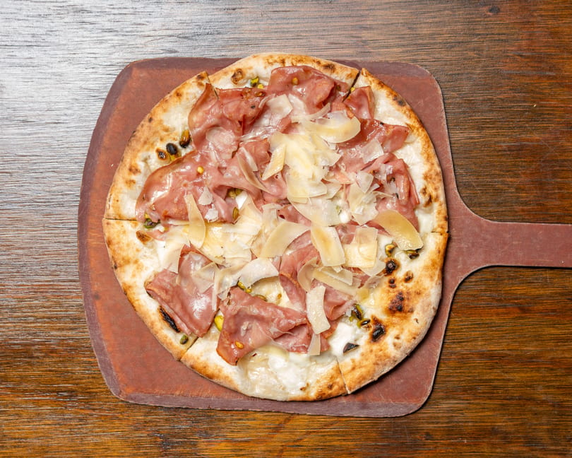A whole pizza with prosciutto, shaved cheese, and pistachios on a wooden pizza peel.
