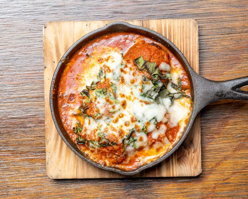 Baked pasta with melted cheese, tomato sauce, and basil in a cast iron skillet on a wooden board.