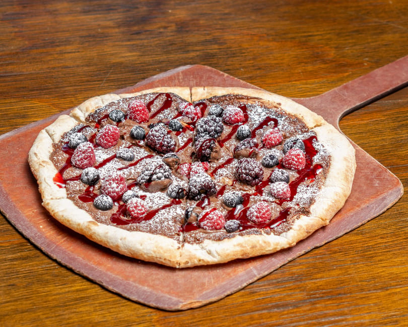 Dessert pizza with chocolate spread, raspberries, blueberries, blackberries, and powdered sugar on a wooden peel.